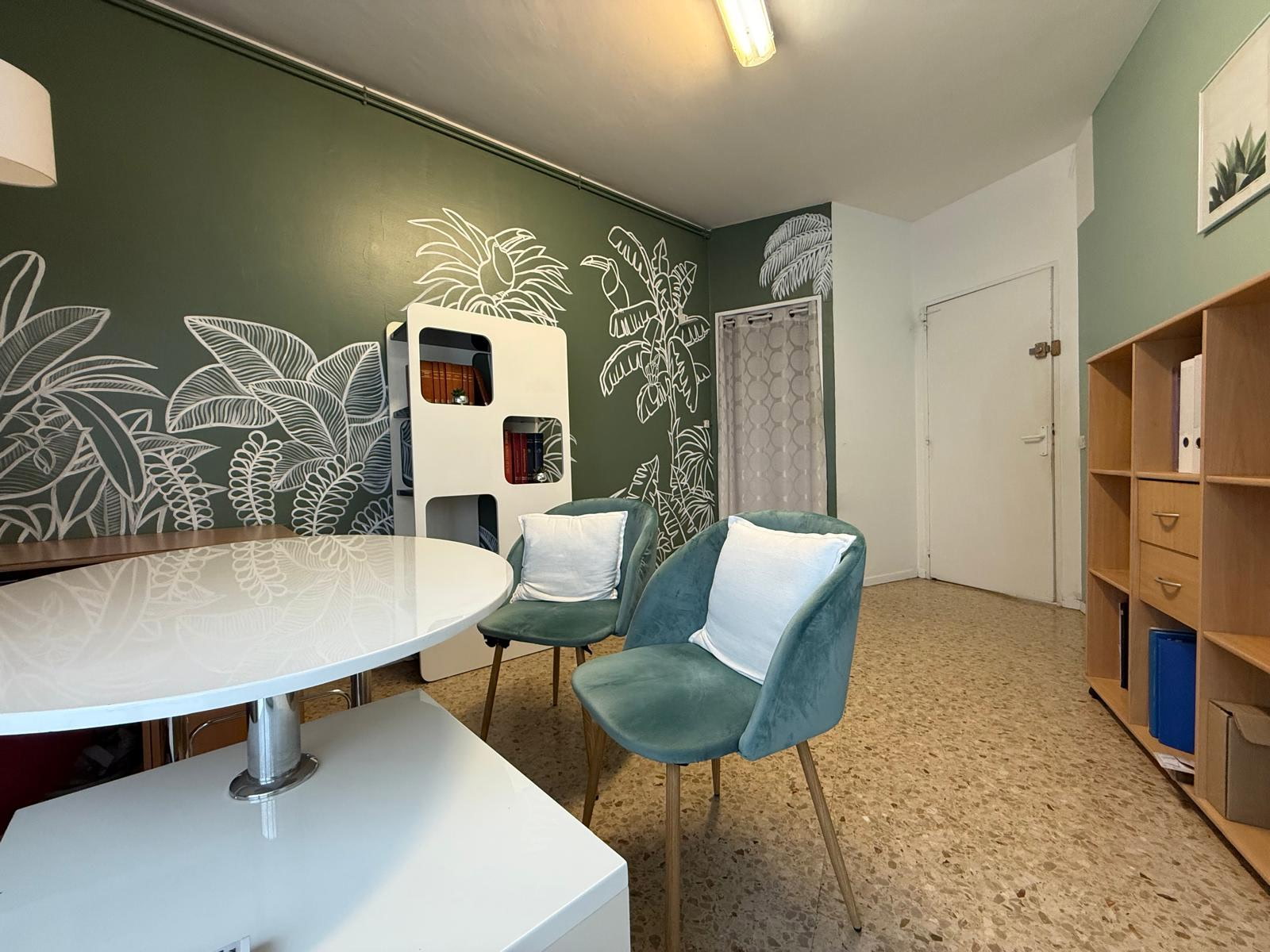 Autre vue du bureau de consultation : les deux fauteuils vert d'eau garnis de coussins blancs, la table ronde et la fresque de feuillages.