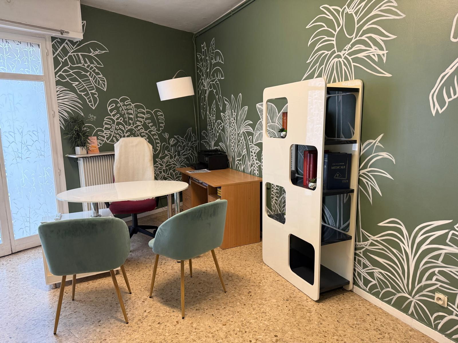 Le bureau de consultation : une table ronde blanche et deux fauteuils en velours vert d'eau, devant un mur vert orné d'une fresque de feuillages tropicaux en trait blanc.