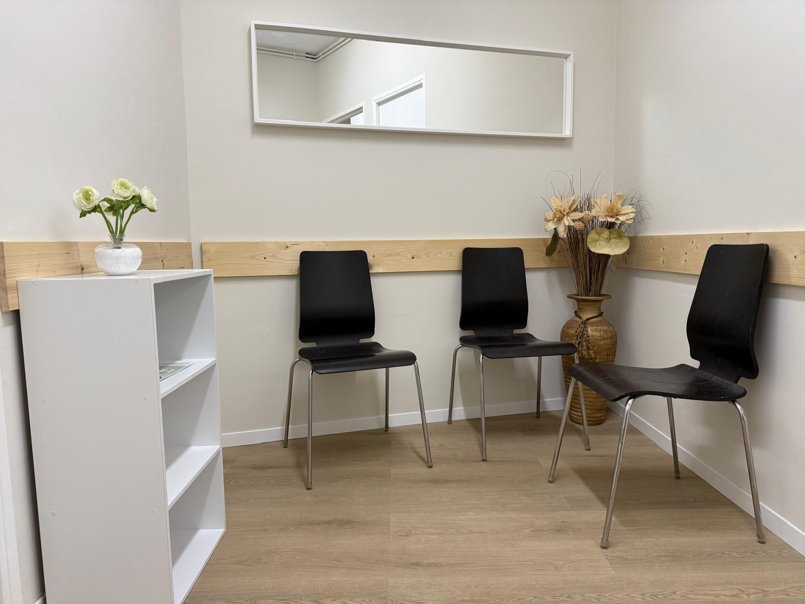 La salle d'attente : trois chaises le long d'un mur clair souligné d'une frise en bois, un miroir et un bouquet de fleurs séchées.